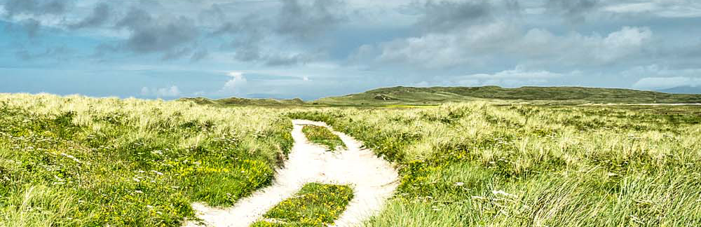 a path cut into sand dunes below a cloudy blue sky on the Island of Uist